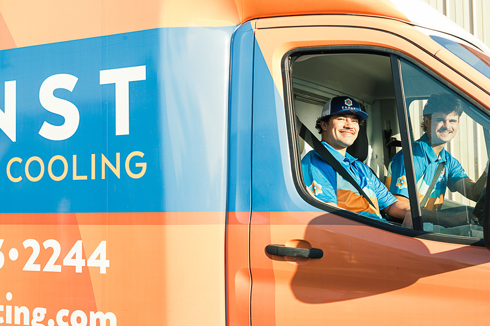 Two uniformed workers smile at the camera from the front seats of an orange and blue service van, seen through the open window.