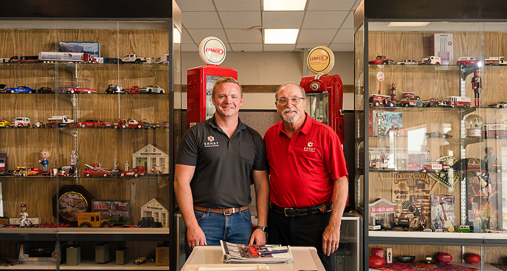 Two men smile as they stand before display cases packed with model cars and memorabilia in a brightly lit room.