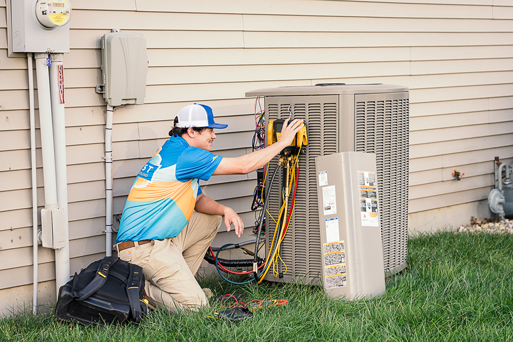 A technician kneels by an outdoor AC unit, actively using diagnostic tools to maintain it, with equipment and a service bag nearby.