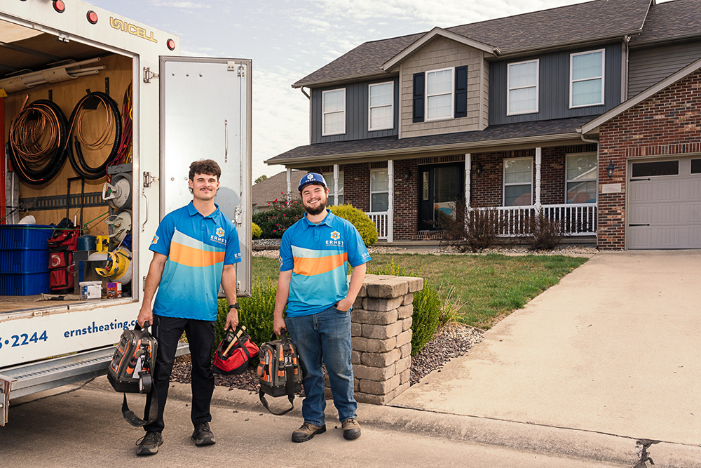 Two HVAC technicians in blue uniforms hold tool bags and smile at the camera beside a service van parked outside a suburban house.